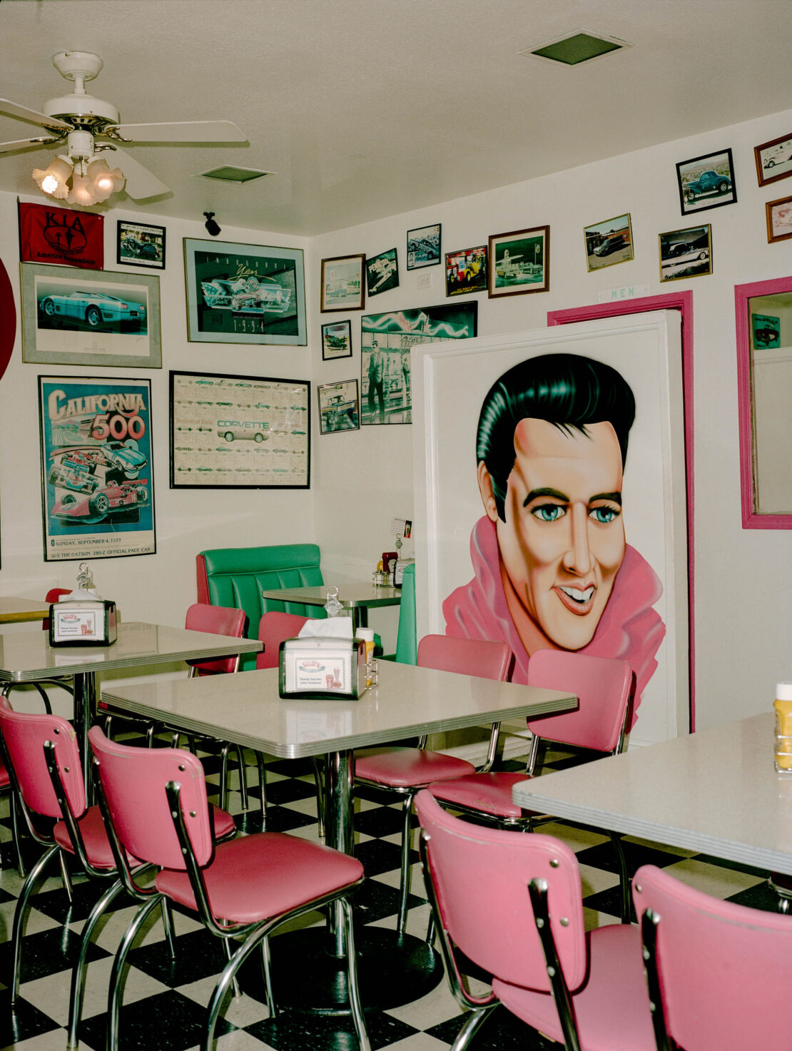Retro diner interior with pink booth chairs, black and white checkered floor, and Elvis Presley art on the wall. Multiple framed car posters.