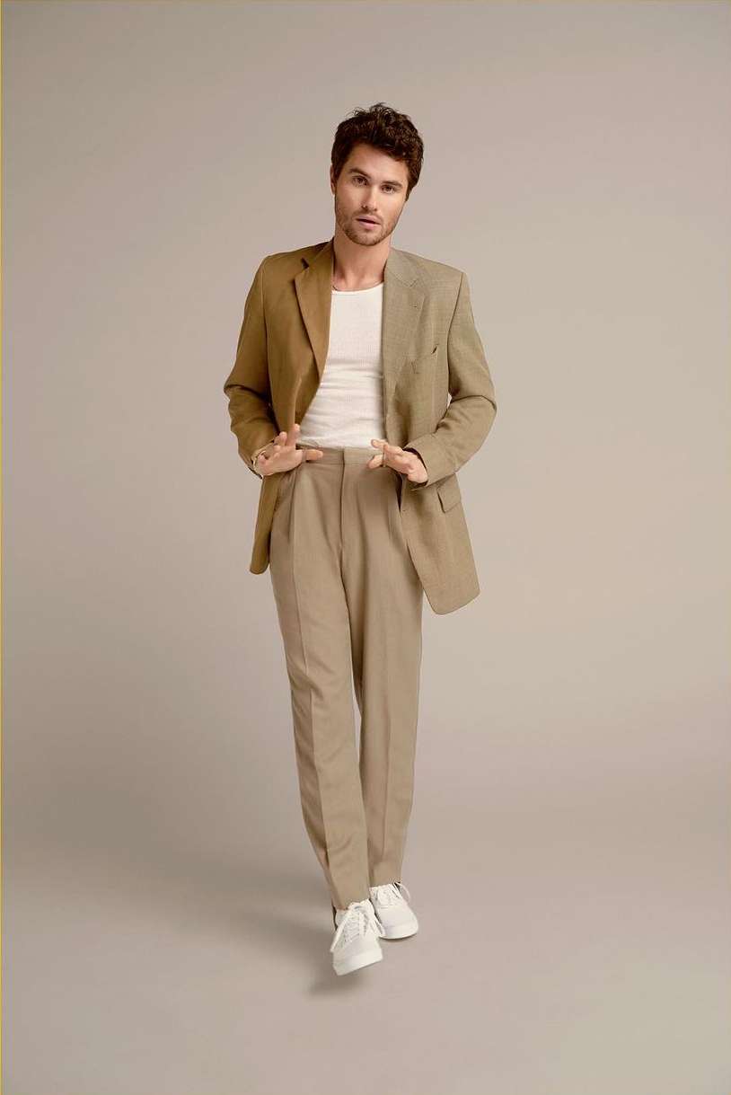 Brunette male model in khaki blazer and pants with white sneakers stands in a full-length shot against a neutral studio backdrop.