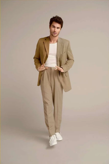 Brunette male model in khaki blazer and pants with white sneakers stands in a full-length shot against a neutral studio backdrop.