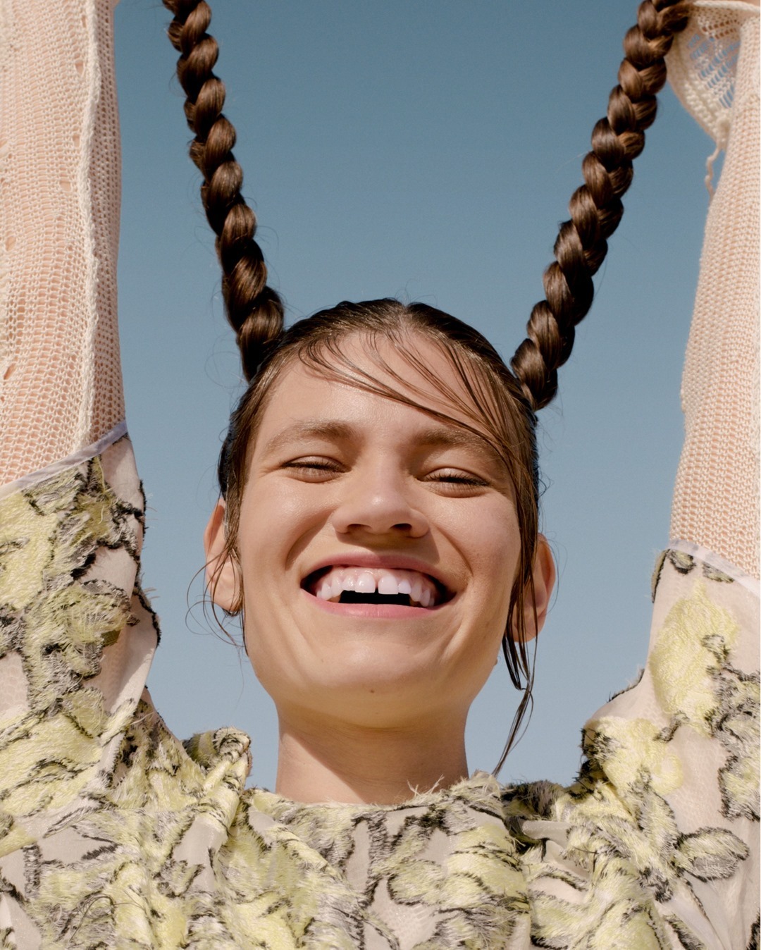 Smiling young woman with braided hair and floral print top poses against a blue sky background in a portrait fashion photograph.