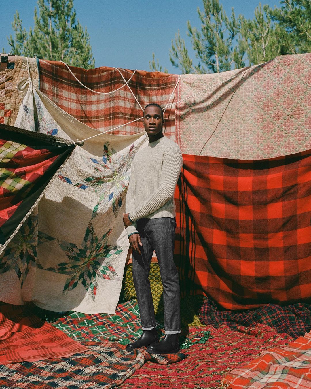 A Black man stands in front of a wall of plaid blankets outdoors. He's wearing a sweater, jeans, and boots, under sunny sky.