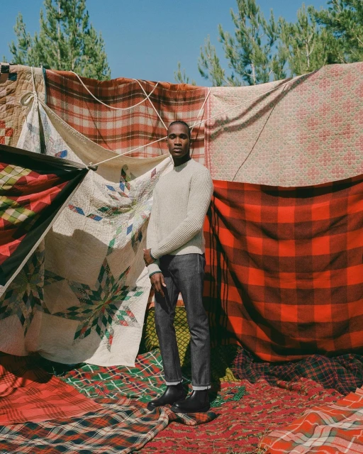 A Black man stands in front of a wall of plaid blankets outdoors. He's wearing a sweater, jeans, and boots, under sunny sky.