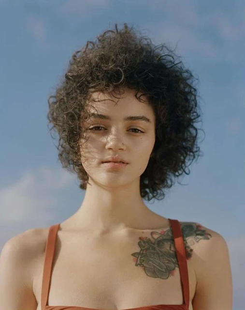 Portrait of a curly-haired woman with a tattoo against a blue sky. The focus is on her face, capturing a natural and serene expression.