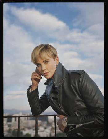 Portrait of a blonde woman wearing a black leather jacket leaning on a railing against a blue sky with clouds, photographed by Amy Harrity