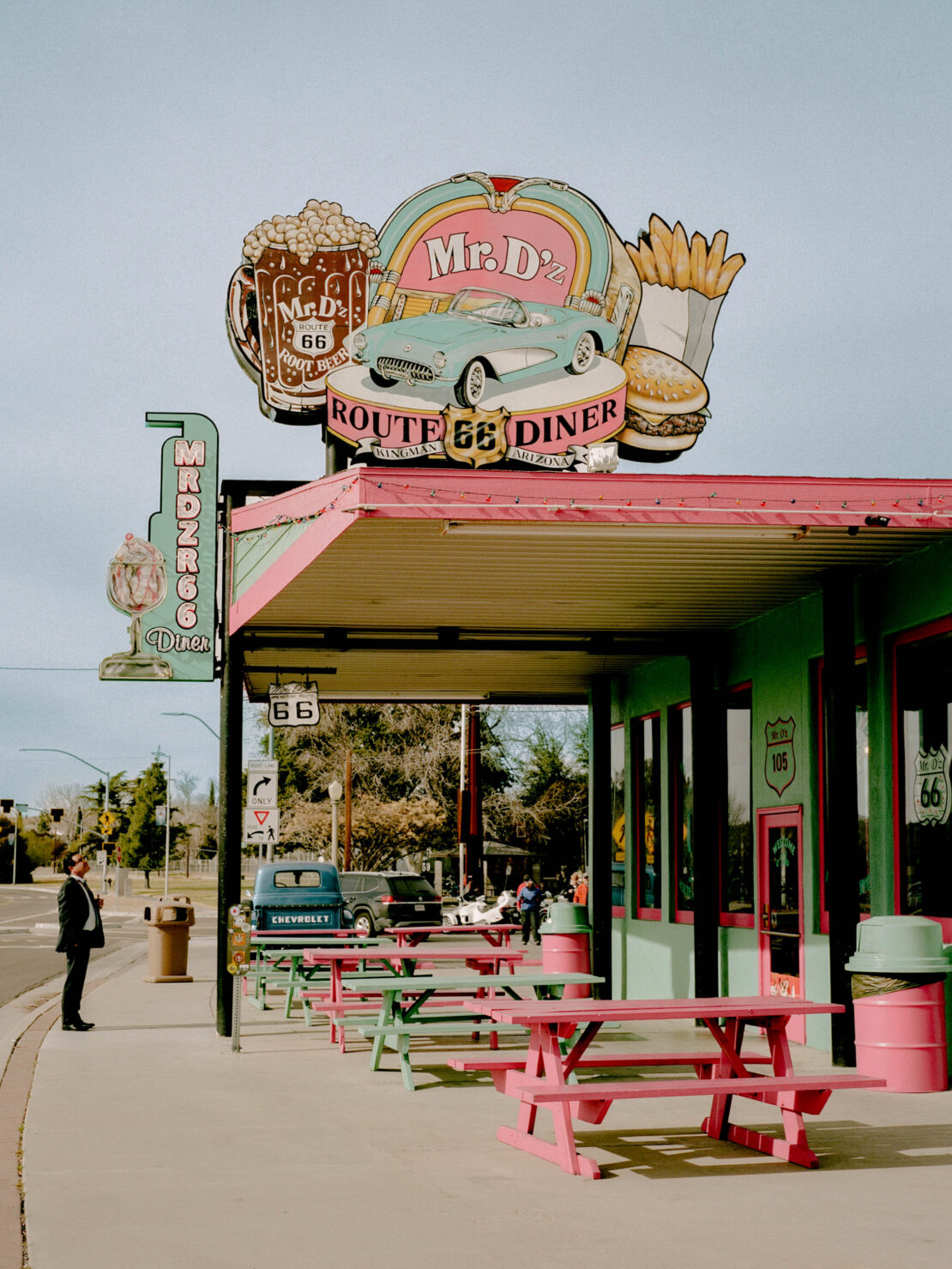 Retro Route 66 diner exterior with vintage signage and classic cars; man in suit standing outside, vintage atmosphere, roadside architecture.