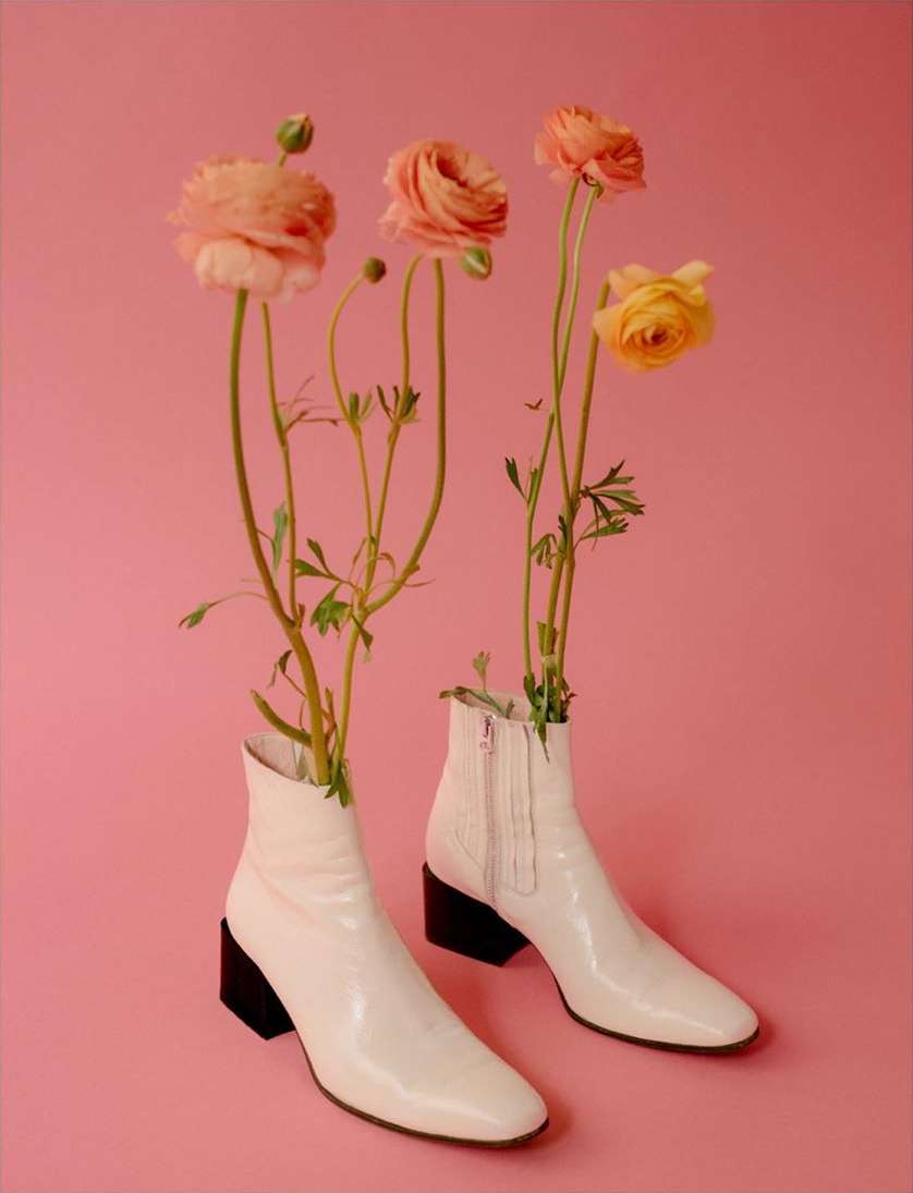 Still life of ranunculus flowers in cream-colored boots against a pink background creating a soft, warm, and delicate visual.