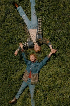 Two people lying on green grass holding hands in an overhead shot, wearing denim jackets and blue jeans.