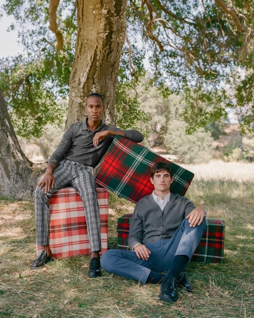 Two adult men pose outdoors in a field next to a tree with gift box props wrapped in red and green plaid fabric.