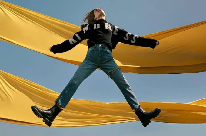 Woman in mid-air wearing jeans, boots and black 'Kindness' sweatshirt with yellow fabric and a clear blue sky in the background.
