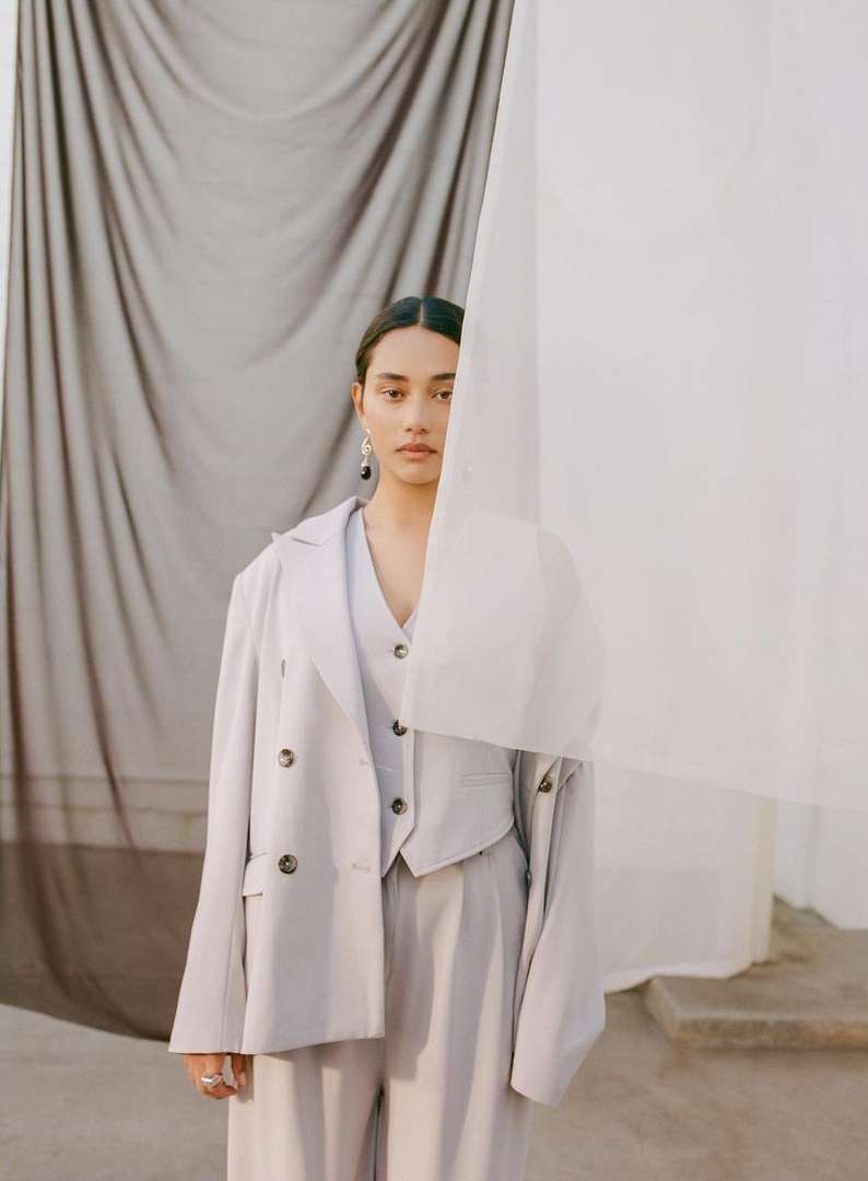Adult female in tailored light grey three-piece suit poses in front of draped grey and white fabrics in a fashion forward portrait.