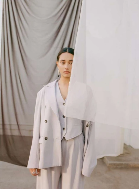 Adult female in tailored light grey three-piece suit poses in front of draped grey and white fabrics in a fashion forward portrait.