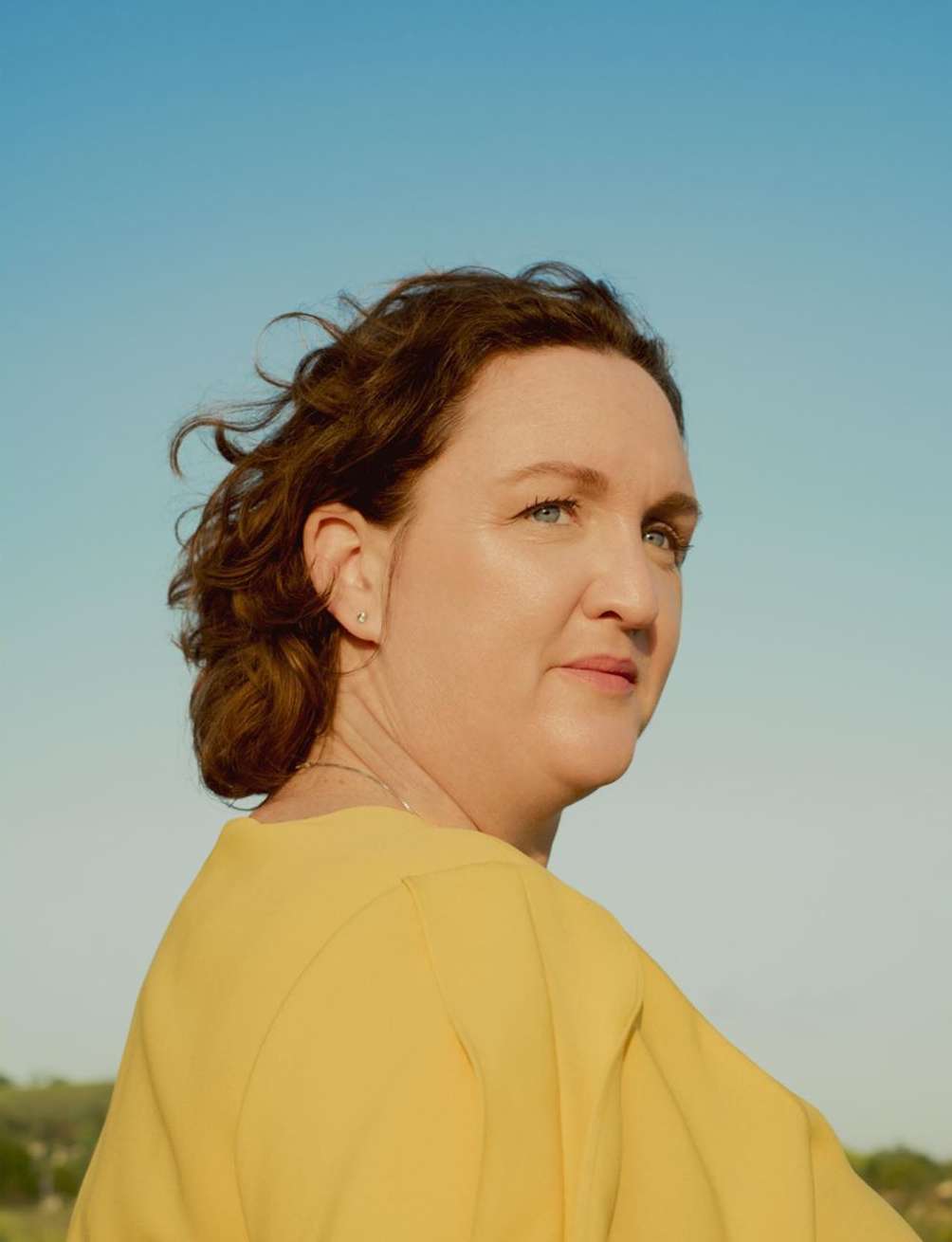 Portrait of a woman with curly brown hair wearing a yellow shirt, set against a clear blue sky and natural outdoor background.