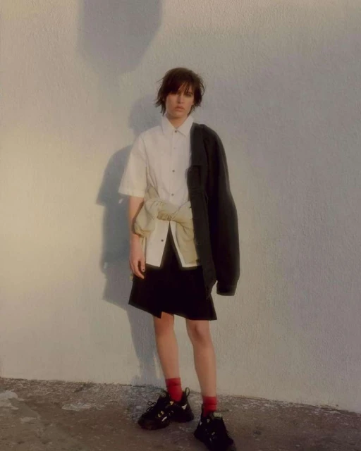 Woman in casual fashion stands against white wall, wearing button-down shirt, dark skirt, black jacket, red socks, and chunky sneakers.