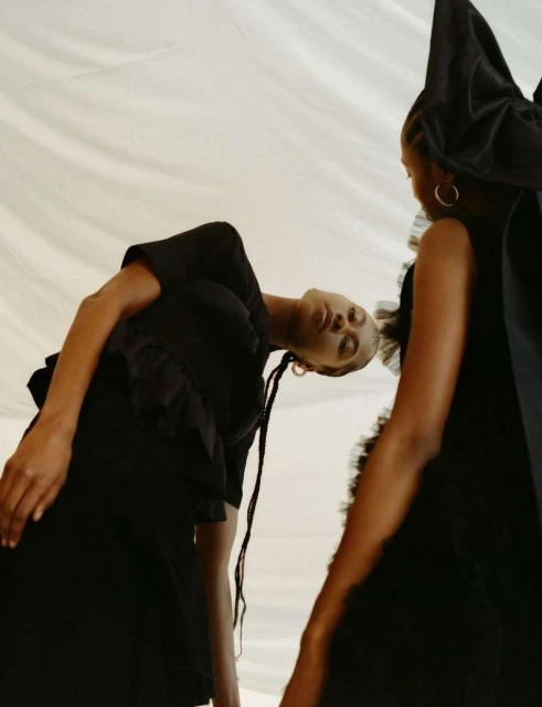 Two Black women pose in black dresses against a white backdrop, showcasing contemporary fashion and style in a studio environment.