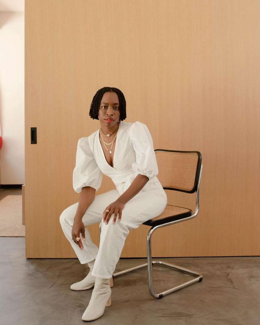 Stylish woman in white seated on a chair indoors. She's wearing a wrap top, pants, and boots against a neutral backdrop, posing confidently.