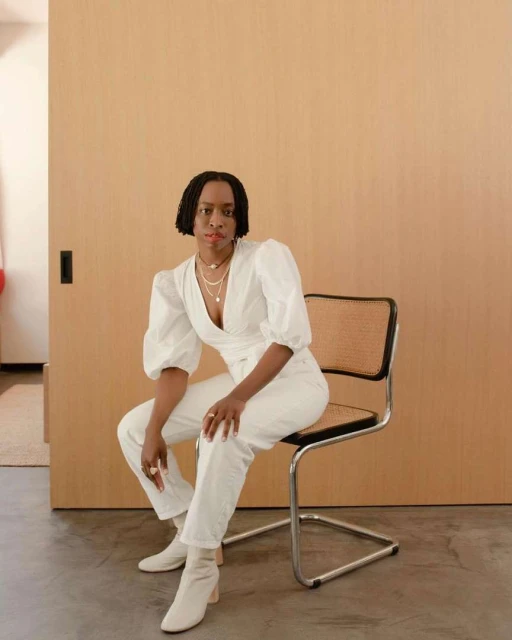 Stylish woman in white seated on a chair indoors. She's wearing a wrap top, pants, and boots against a neutral backdrop, posing confidently.