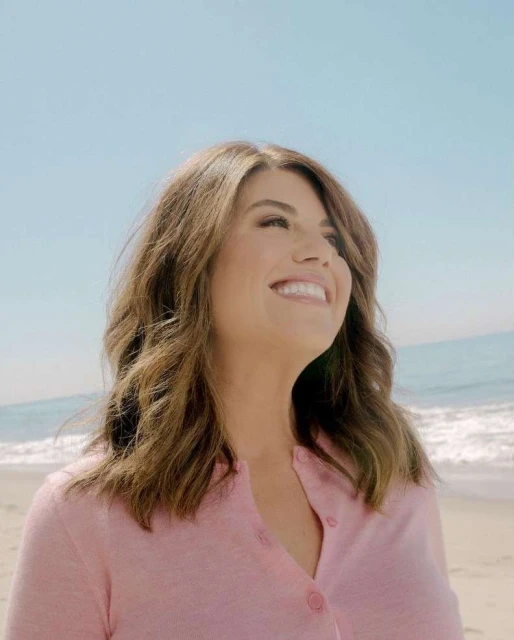 Smiling adult woman with shoulder-length brown hair in a pink cardigan on a sunny beach with a clear blue sky.