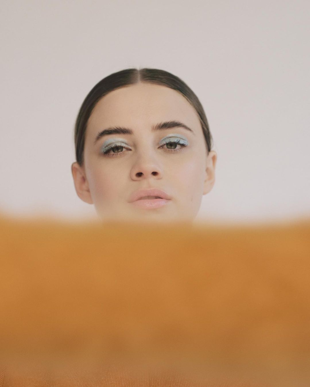 Close-up portrait of a woman with light blue eyeshadow and a neutral facial expression against a white and tan backdrop.