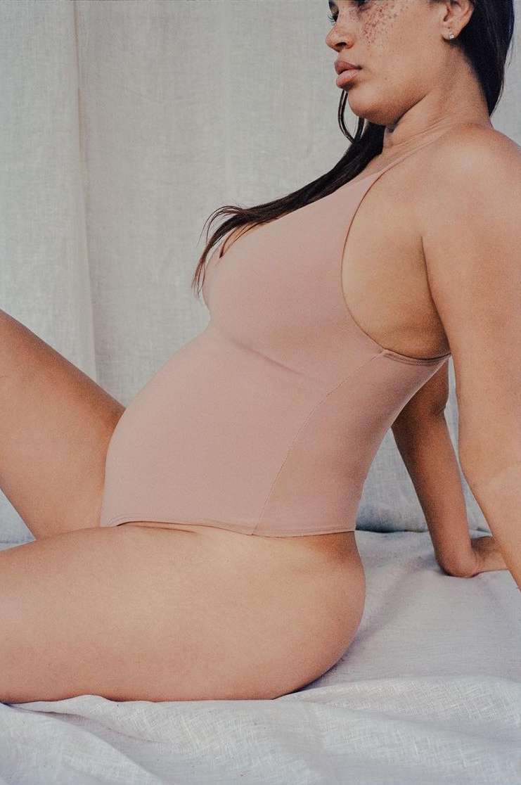 A woman poses in a close-up profile shot wearing a neutral-toned bodysuit against a simple background, showcasing body positivity.