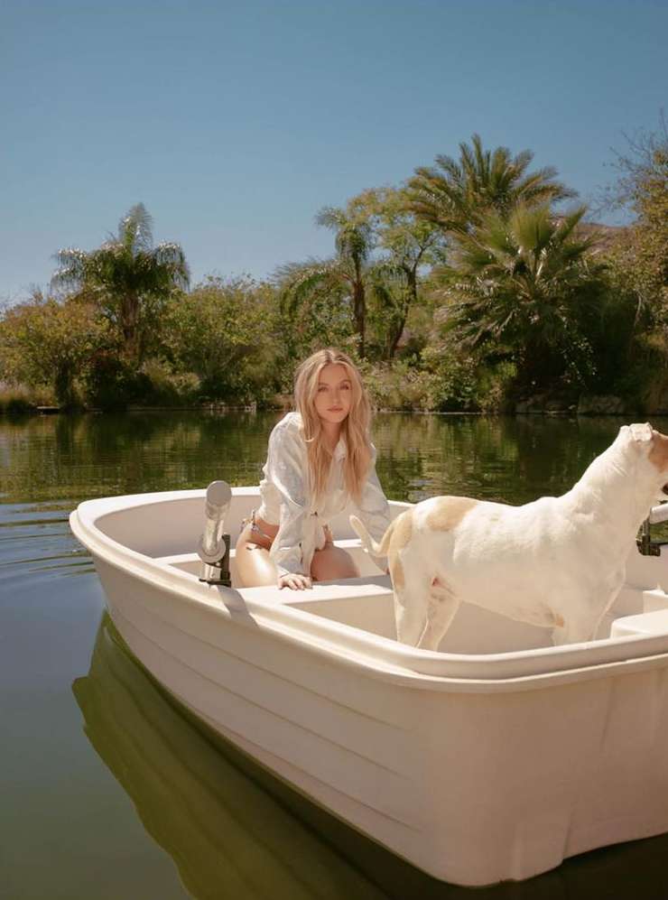 Woman and white dog in a rowboat on a sunny day. Trees and clear blue sky in background. Woman wears a white shirt. Still water.