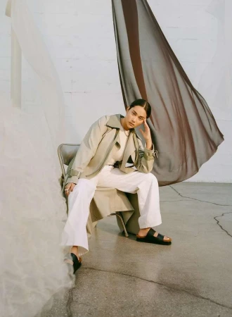 Model seated on a chair in a trench coat and white trousers with hanging sheer fabric draped in the background