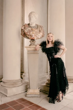Elegant woman in a black gown poses beside a marble bust in an indoor setting with large columns and a tiled floor.