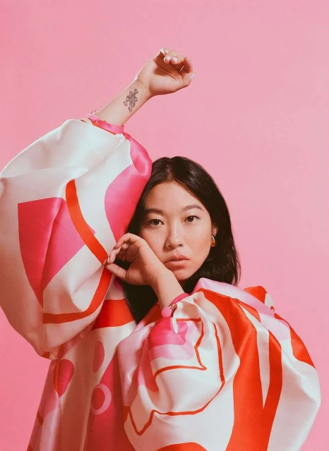 Eye-level portrait of a model with dark hair in an oversized patterned top against a pink background, posing with a raised arm and hand near face.