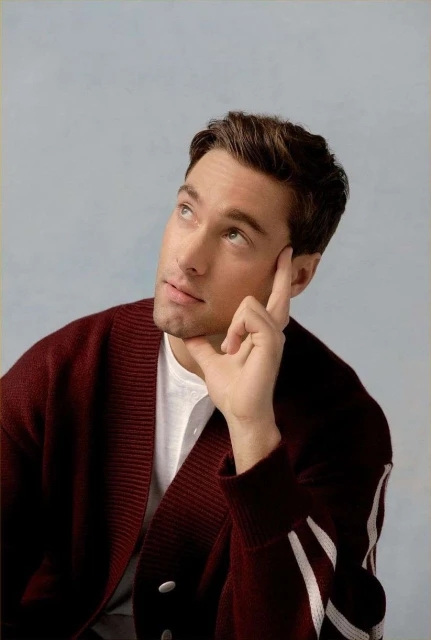 Portrait of a fair-skinned adult man with brown hair looking up and resting his face on his hand while wearing a burgundy cardigan.