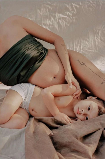 Mother and child lying together on a bed. Woman with bare stomach is draped in green fabric, child is in white shorts on beige sheet.