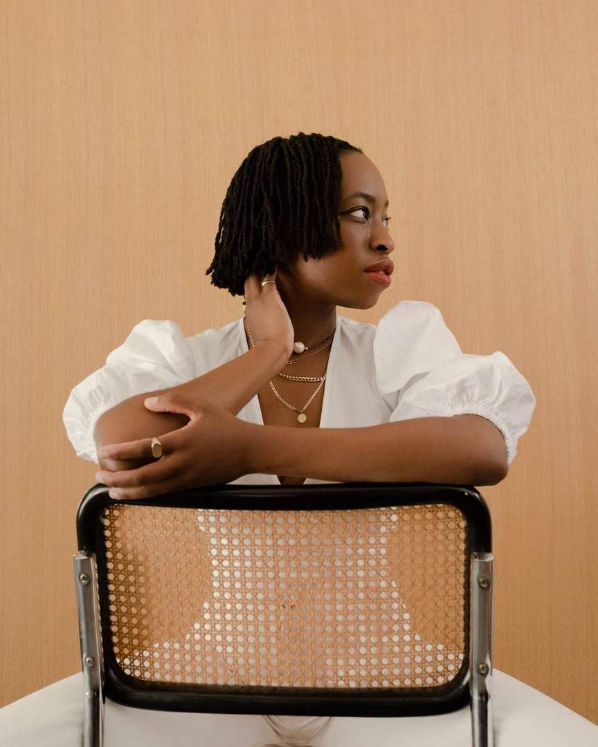 Portrait of Black woman with short locs in white blouse, seated on cane chair against neutral backdrop, gazing off-camera.