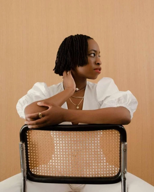 Portrait of Black woman with short locs in white blouse, seated on cane chair against neutral backdrop, gazing off-camera.
