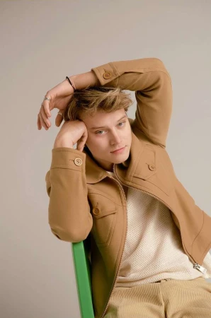 Young man in a camel jacket and white top sitting on a green chair against a plain studio background.