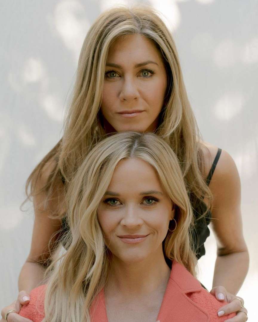 Jennifer Aniston and Reese Witherspoon pose together in a portrait with a light backdrop and natural lighting, both smiling slightly.