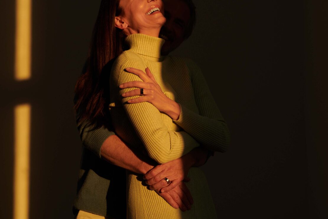 Warm embrace: A woman in a lime dress is embraced by a person in a turquoise sweater, bathed in light with a dark background.