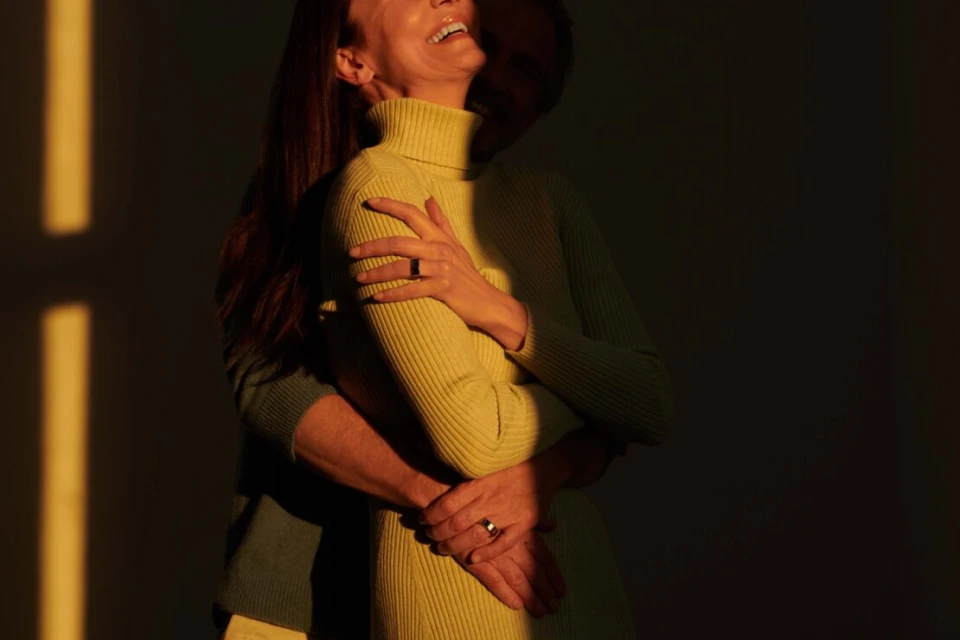 Warm embrace: A woman in a lime dress is embraced by a person in a turquoise sweater, bathed in light with a dark background.