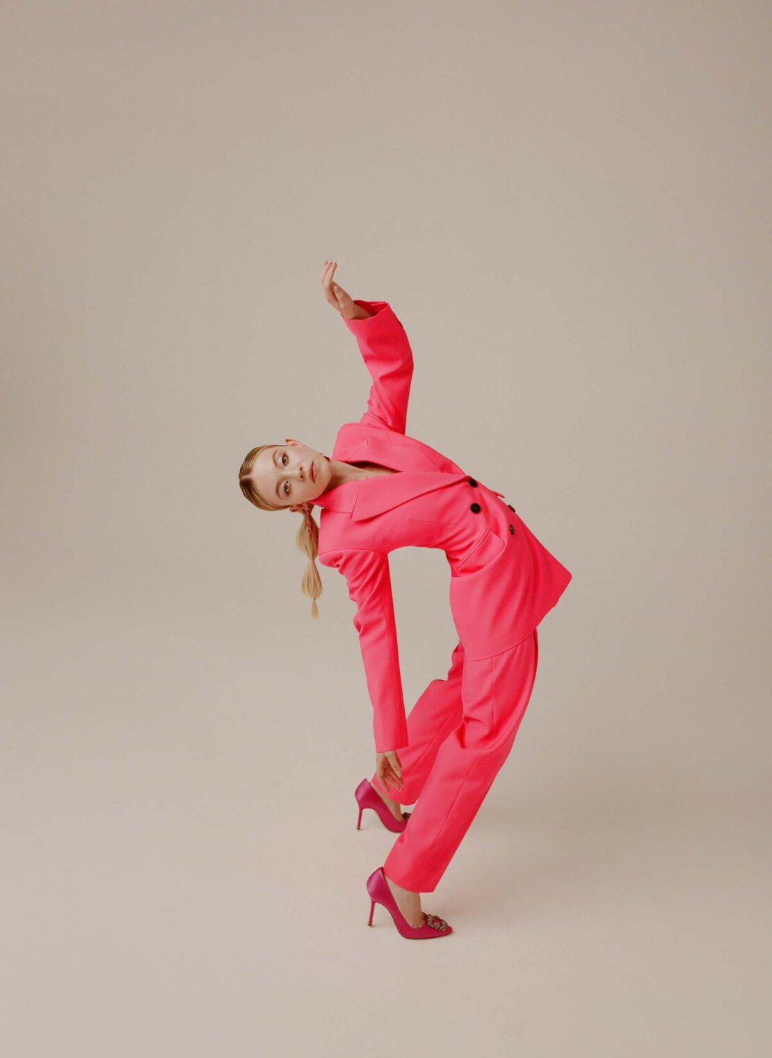Woman in vibrant pink pantsuit and matching heels arching backward against a seamless light grey studio backdrop.