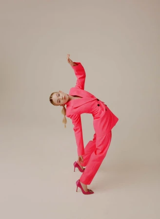 Woman in vibrant pink pantsuit and matching heels arching backward against a seamless light grey studio backdrop.