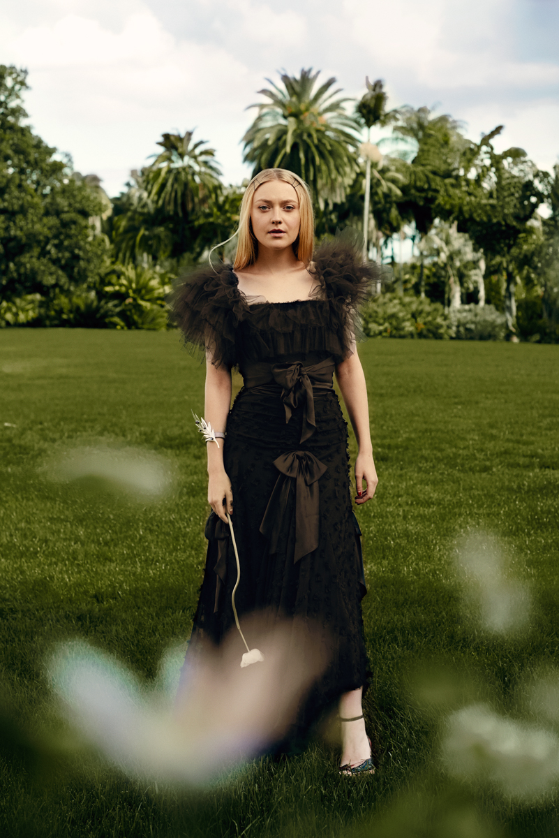 Dakota Fanning in black gown poses outdoors on green grass near palm trees, holding a flower, fashion photography, beauty editorial.