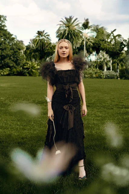 Dakota Fanning in black gown poses outdoors on green grass near palm trees, holding a flower, fashion photography, beauty editorial.