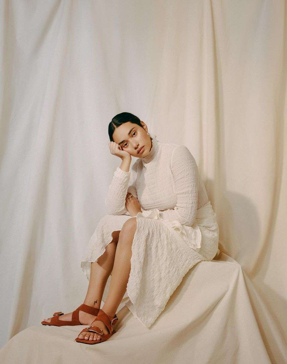 A young woman in white textured dress and brown sandals sits on draped fabric in a photo studio with soft, neutral lighting.