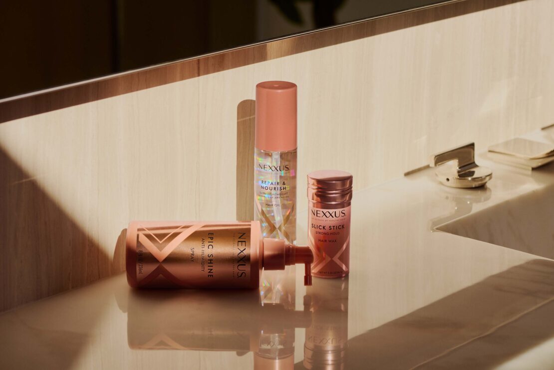 Three Nexxus hair product bottles, including Epic Shine anti-humidity spray and Slick Stick hair wax, displayed indoors on a reflective surface.