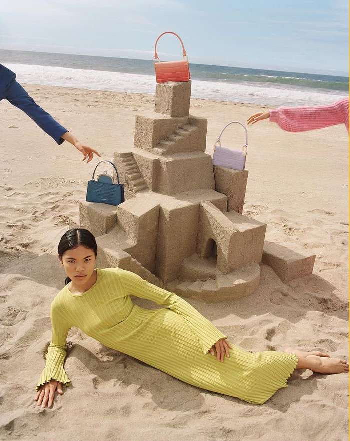 A woman in a green dress lays on a sandy beach next to a sandcastle holding colorful handbags, with arms reaching toward the bags.