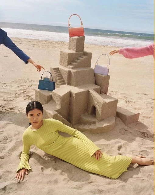 A woman in a green dress lays on a sandy beach next to a sandcastle holding colorful handbags, with arms reaching toward the bags.