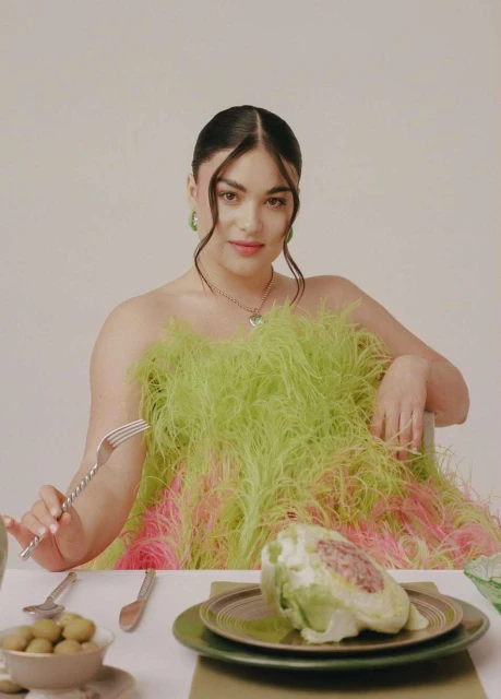 Brunette woman in an elaborate feathered dress sits at a table with plates, silverware, and food, including a head of lettuce.