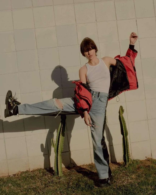 A person in a white tank top, ripped blue jeans, and black shoes poses with a red jacket on a cactus against a white block wall outdoors.