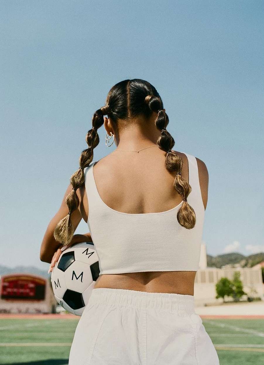 A woman with braided hair holding a soccer ball on a sports field. She is wearing a white top and shorts, standing outside.