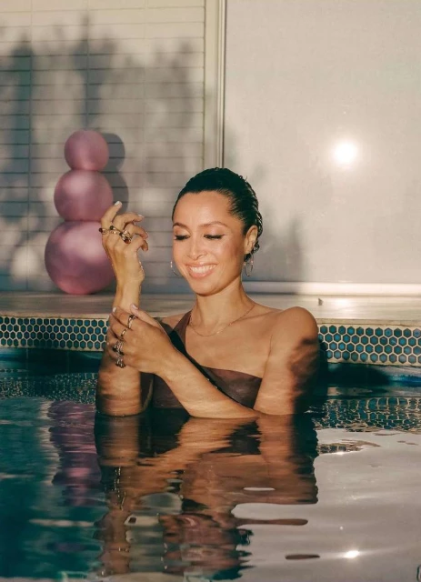 Smiling woman in a brown swimsuit, posing in a pool with her hand up, wearing rings, with a large pink sculpture in the background.