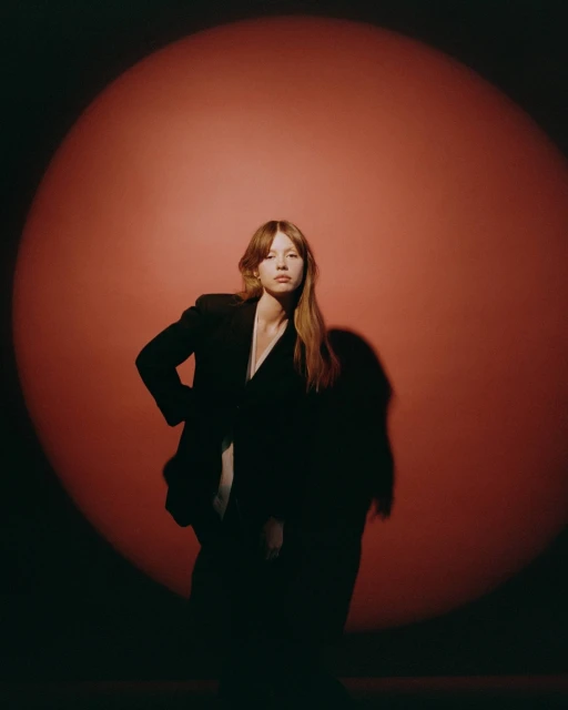 Portrait of an adult female posing in a dark blazer against a red backdrop with spotlight, creating a dramatic, high-contrast studio shot.