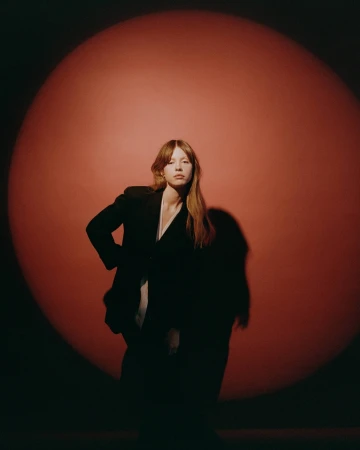 Portrait of an adult female posing in a dark blazer against a red backdrop with spotlight, creating a dramatic, high-contrast studio shot.