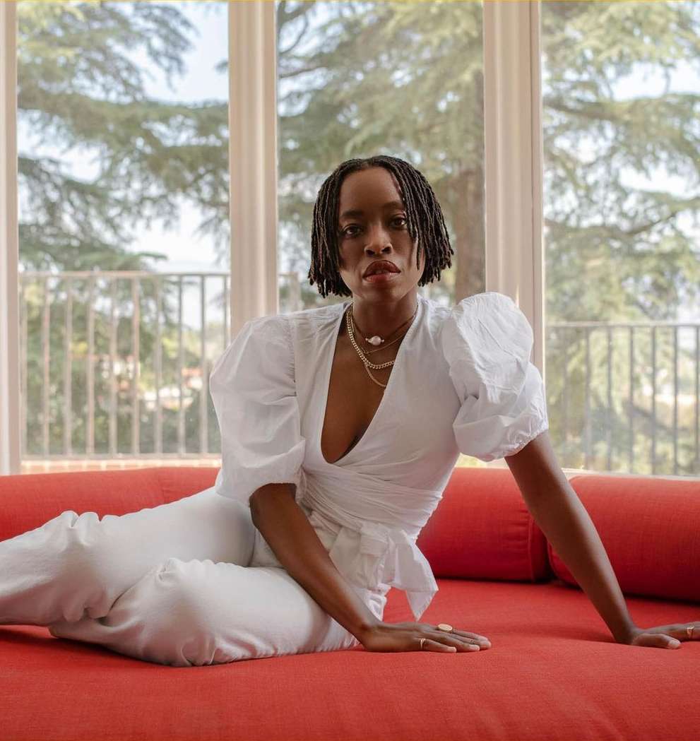 Stylish woman in white outfit poses on orange couch in sunlit room with large windows. She wears layered necklaces and subtle makeup.